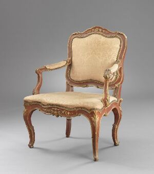  An antique wooden armchair with curved arms and intricately carved golden-brown frame, upholstered in a faded beige fabric, against a neutral gray background.