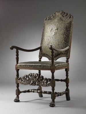  An antique, intricately carved wooden chair with faded upholstery on the seat and high back, standing against a soft gray background. The wood is a deep brown color, and the fabric appears to be a worn pale taupe or gray, indicative of historical use and age.