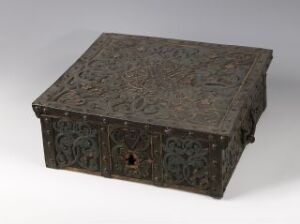  An antique dark brown or black carved wooden or metal box with intricate floral and vine-like relief patterns throughout, featuring a keyhole in the front, set against a neutral background, suggesting age and craftsmanship.