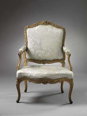  An antique gilded chair with creamy white upholstery, featuring a curved backrest with intricate carvings and decorative motifs. The chair's arms and cabriole legs are also adorned with gold leaf and carving, suggesting a baroque or rococo style.