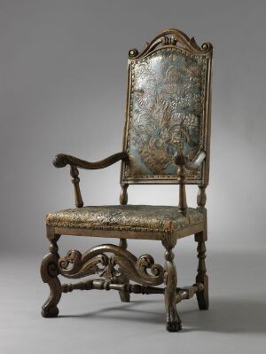  An antique, ornately carved armchair with faded floral upholstery in muted blues, greens, and cream, set against a plain gray background. The dark wooden frame features intricate baroque-style carvings, and the chair's pattern shows signs of age and use.