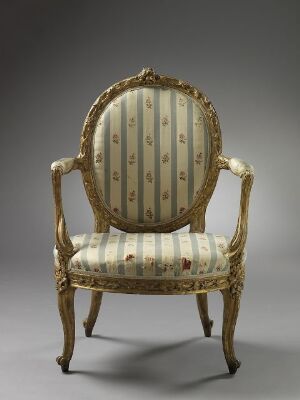  An opulent armchair with gilded wooden frame featuring intricate carvings and upholstered with a striped, floral-patterned fabric in muted tones, set against a simple gray background.