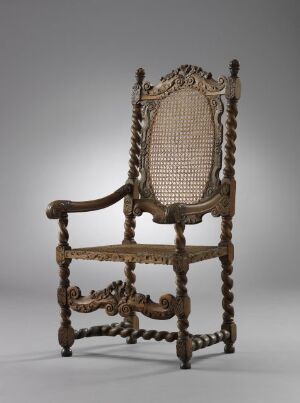  An antique baroque chair with a high, oval cane backrest framed with intricate carvings, barley twist posts and legs, and ornate scroll-shaped armrests against a neutral gray background.