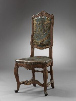  An antique wooden chair with high backrest, intricately carved and upholstered with a patterned fabric in shades of green with gold, beige, red, and blue accents. The chair exhibits a baroque or rococo style and is presented against a neutral gray background.