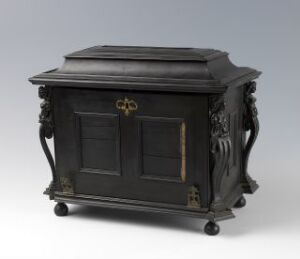  Antique black wooden cabinet with sloped top, ornate foliage carvings on the sides, brass loop handles on the doors, and set on small rounded feet. The doors are accented with a thin golden-brown trim.