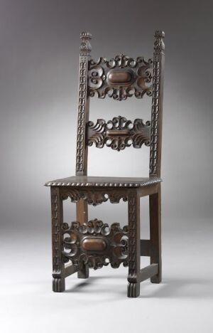  An ornate dark wooden chair with elaborate openwork carvings on the backrest, featuring a raised central medallion and a lattice-like design with geometric and floral motifs, set against a plain grey background.