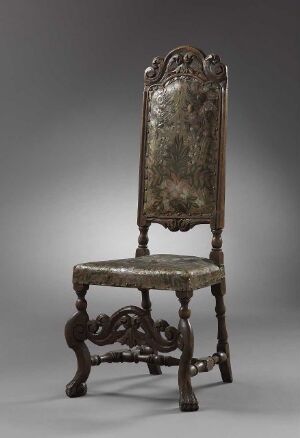  An antique wooden chair with a high, scrolled backrest featuring baroque-style carvings. The chair has ornate upholstery in faded silver-gray, and the dark wooden legs are joined by a curved, carved stretcher.