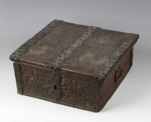  An antique, ornately carved metal chest with a patinated grey surface showing signs of age and featuring sturdy bands with rivets, a prominent lock, and a side handle, against a light backdrop. Artist name and title are unknown.