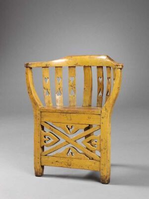  An antique wooden chair with mustard-yellow paint and decorative X-shaped cut-out patterns, set against a soft grey background. The paint shows signs of wear, emphasizing the chair’s age and history.