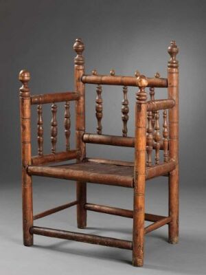  An antique wooden armchair with ornately turned front legs and spindles, featuring a warm brown color and detailed wood grain, set against a simple grey background. The chair has a flat wooden seat, low armrests, and two decorative turned finials atop the backrest.