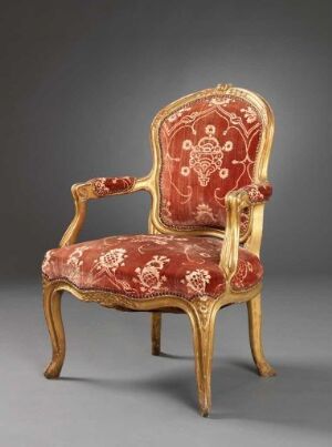  A vintage armchair with intricate light-colored wooden frames and plush red upholstery featuring floral patterns and gold-colored designs, set against a simple gray background.