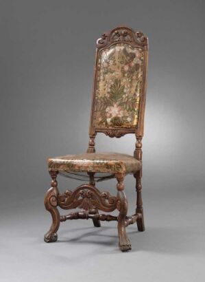  An antique wooden chair with a high backrest and seat upholstered in a floral tapestry fabric, surrounded by intricate wooden carvings in a dark brown finish, set against a neutral gray background.