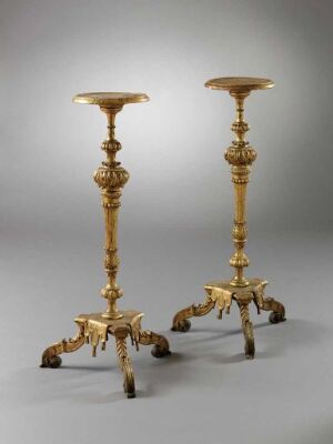  A pair of ornate, tall candlestick holders with a golden finish, presenting a tripod base with decorative animal paw-like feet, intricately designed stems featuring nodes and floral motifs, culminating in a shallow dish at the top, set against a neutral light grey background.