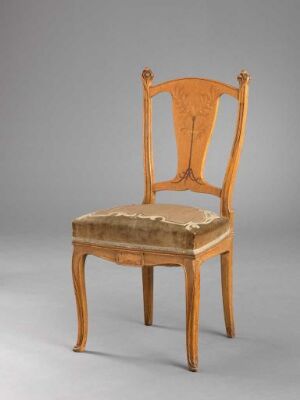  An antique wooden chair with a honey-colored frame and a faded beige upholstered seat, set against a plain gray background. The backrest has an ornate lyre-shaped splat design, and the chair exhibits signs of age and usage.