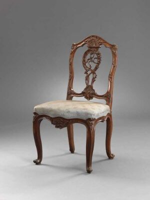  Antique wooden chair with curvy legs and a cream cushioned seat, featuring a decorative, symmetrical backrest with intricate wood carvings against a plain gray background.