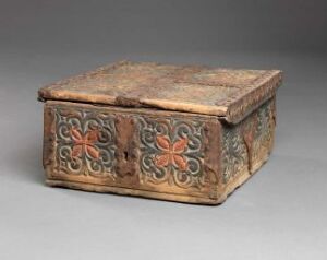  An old metal box with a hinged lid, featuring rusted brown tones and red floral motifs on its sides, displayed on a grey background. Artistname and title are unknown.