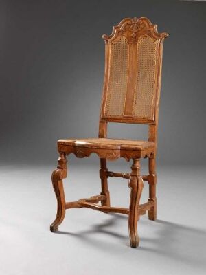  An antique wooden chair with ornate carvings, standing on a grey floor against a dark, gradually lightening background. The chair features a high backrest with a caned panel and a matching caned seat, supported by turned front legs and simpler back legs, connected by stretchers.