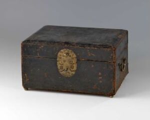  An antique wooden box covered in dark brown leather with scuffs and signs of wear, featuring a tarnished golden coat of arms on the lid, set against a neutral grey backdrop.