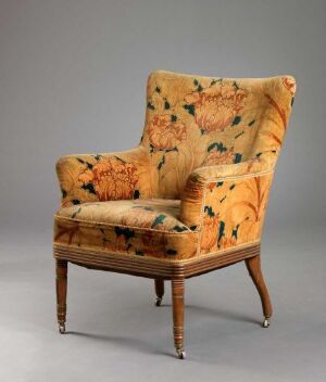  Antique floral upholstered armchair with wooden legs and a woven wicker section at the base, set against a neutral gray background.