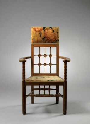  An antique wooden chair with a floral motif on the upper back panel and seat, featuring a tall back with geometric cut-out patterns, sturdy armrests, and dual-level stretchers connecting the legs. It stands against a neutral gray background.