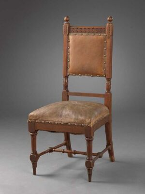  An antique wooden chair with faded tan leather upholstery on the seat and backrest, featuring studded detailing and turned wood legs, presented against a simple gray background.