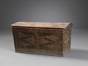  A weathered rectangular wooden chest with a slightly domed lid and a series of diagonal carvings creating a diamond pattern on the front, set against a gradient gray background.