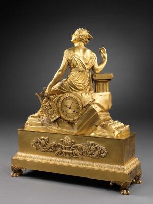  An ornate gold-toned mantel clock with a classical figurative sculpture of a draped woman alongside a clock face with Roman numerals, set on a decorative base with relief motifs, against a dark gray background. Artist and title unknown.