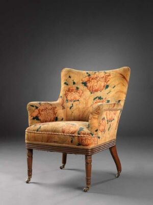  An antique armchair with beige upholstery featuring large orange and rust-colored floral patterns, set against a soft gray background with the wooden details of the armrests and legs finished in a dark brown tone.