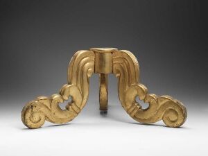  An ornate golden decorative stand or furniture mount with symmetrical baroque-style volutes and scrolls, set against a neutral grey background.