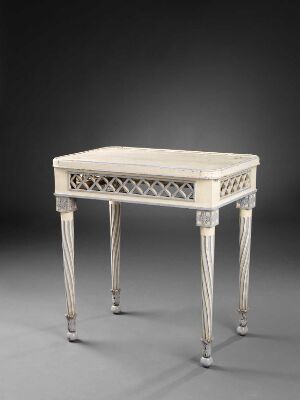  An elegant, antique neoclassical table with a creamy white tabletop, intricate trefoil patterned edges, and tapered legs ending in metallic feet, against a neutral gray backdrop.