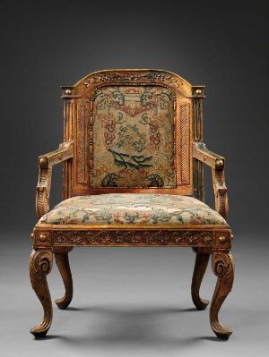  An ornate wooden armchair with cabriole legs and upholstered with a tapestry-style fabric featuring a detailed peacock design on the backrest and floral patterns on the seat, set against a dark background.