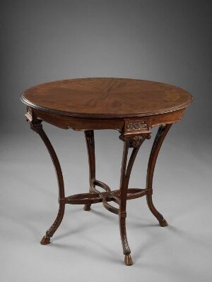  An antique round wooden table with a polished brown top, intricately detailed legs, and a smaller central shelf, set against a neutral gray background. Artistname and title are unknown.