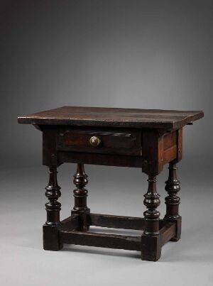  An antique dark brown wooden table with turned legs and a single drawer with a round knob, set against a graduated grey background.