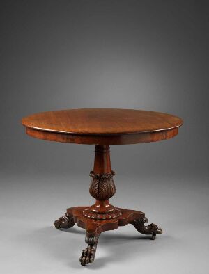  An antique circular wooden table with a detailed, turned column and ornate legs on a neutral, dark-grey background. The table displays intricate carvings and a polished, rich amber surface.