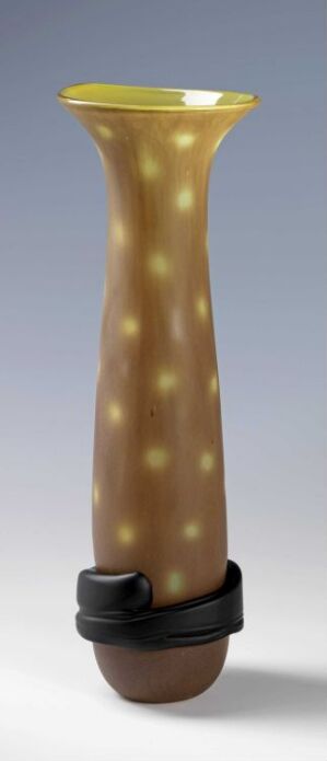  "Åpent frø med spor," a blown glass artwork by Ulla-Mari Brantenberg, features a tall, seed-like structure that transitions from earthy brown to tan with yellow polka dots and a bright yellow rim, cradled by a dark ring at the base.