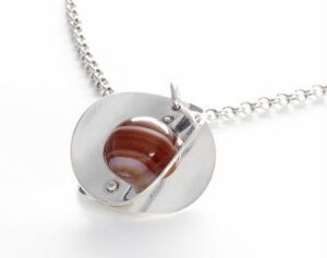  A silver necklace with a circular design featuring a red marbled stone set within the silver disc, part of Tone Vigeland's "Punch" series, reflecting simple elegance through its silver construction and earth-toned gemstone centerpiece.