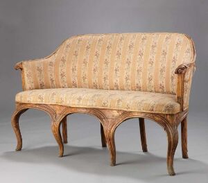  An antique-style wooden settee with a curved back and arms, featuring a light wood frame with a prominent grain and upholstered with a cream-colored fabric adorned with pink and green floral stripes.