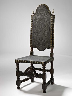  An antique wooden chair with intricate carvings and dark upholstery, adorned with metallic studs, standing against a light gray background.