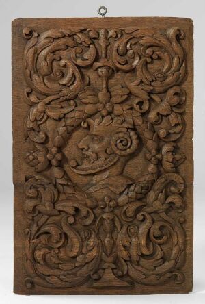  A rectangular wooden panel with elaborate symmetrical acanthus leaf and scrollwork carvings centered around a vase-like figure, displaying a range of warm brown tones with a small metal ring on the top center for hanging.