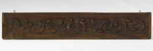  An antique, dark brown, wooden panel with symmetrical, intricately carved floral and foliage motifs, mounted on a wall with metallic fixtures at either end.