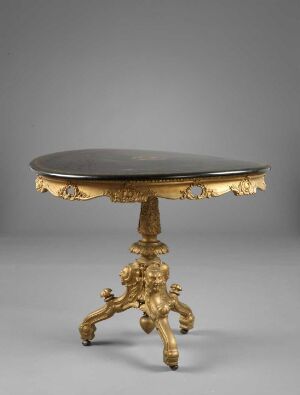  An opulent circular table with a dark top and a lavishly decorated golden base featuring intricate designs and curved legs set against a neutral gray background.