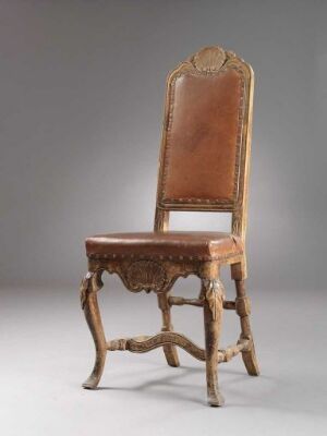  An antique chair with a high, curved backrest upholstered in caramel-brown leather and a wooden frame with dark brown ornate carvings. The chair stands against a plain gray backdrop.