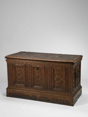  An antique dark brown wooden chest with intricate carvings on the front and sides. The chest features a flat top and stands prominently against a plain light grey background, highlighting the detailed craftsmanship.