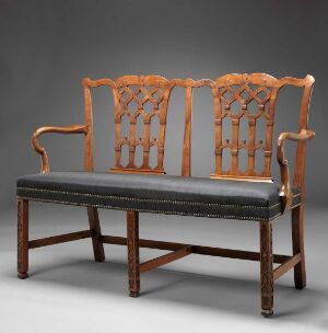  An antique wooden double-seated chair with elaborate open-work back design, finished in a warm brown tone, and upholstered with dark leather on the seat, set against a plain dark gray background.