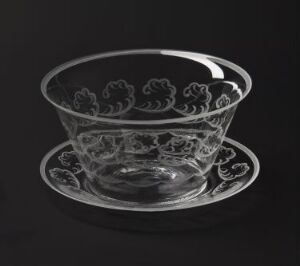  A clear glass bowl with engraved floral designs by Orrefors Glasbruk A/B, resting on a matching glass plate against a dark background.