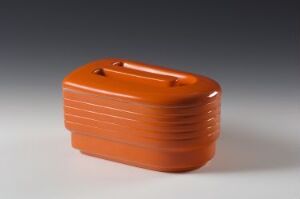  Small, rectangular, terracotta-orange object with rounded edges and horizontal ridges, featuring a coin slot on top, against a gradient grey background. Artistname and title are unknown.