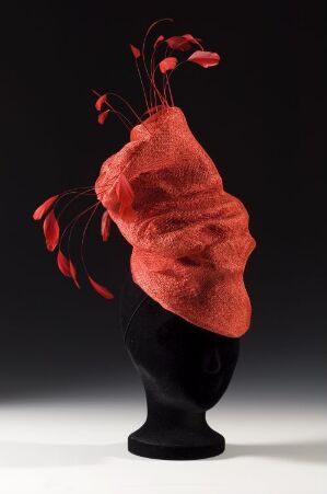  Abstract sculpture featuring a twisty, deep red porous form with long, thin black tendrils topped with darker red buds displayed on a black stand against a dark background.