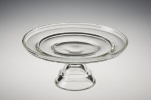  Transparent glass cake stand with raised edges and a concentric circular pattern, made by Sverre Pettersen, on a background gradient from white to light gray.