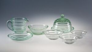  A glassware set titled "Sola" by artist Sverre Pettersen, featuring translucent green-tinted pressed glass objects including a pitcher, nested bowls, a large serving plate, and a dome-covered butter dish with a concentric circle design, arranged against a gray to white gradient background.