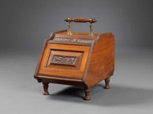  Antique wooden box with a sloped top and decorative carved front panel, fitted with a brass handle and small legs, displayed against a grey backdrop.